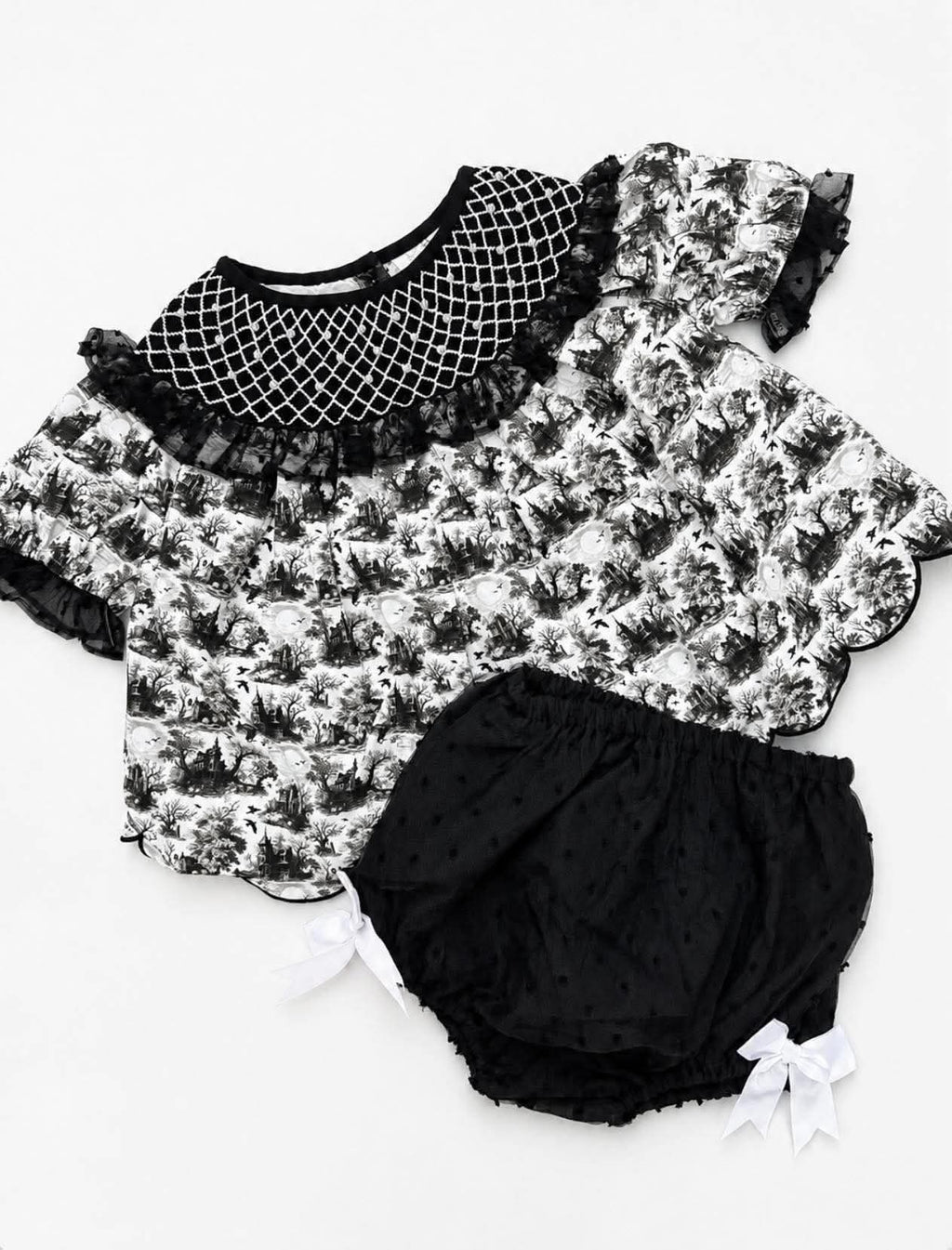 A baby girl's two-piece outfit set featuring a short-sleeve top with a black and white toile floral print, a black fishnet collar with ruffles, and matching black polka-dot bloomers with white bow details, laid flat on a white background.