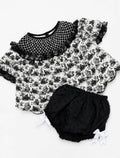 A baby girl's two-piece outfit set featuring a short-sleeve top with a black and white toile floral print, a black fishnet collar with ruffles, and matching black polka-dot bloomers with white bow details, laid flat on a white background.